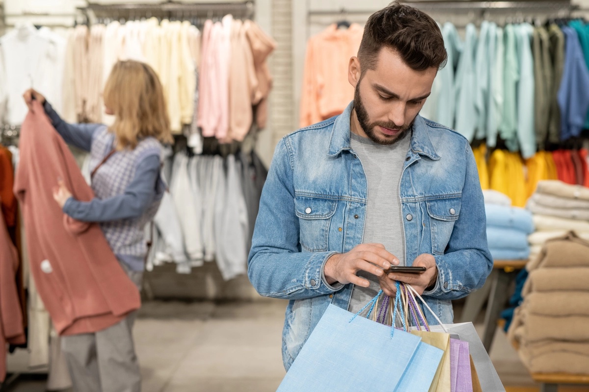 A man with a shopping addiction on his phone while standing in a retail store and holding multiple bags.