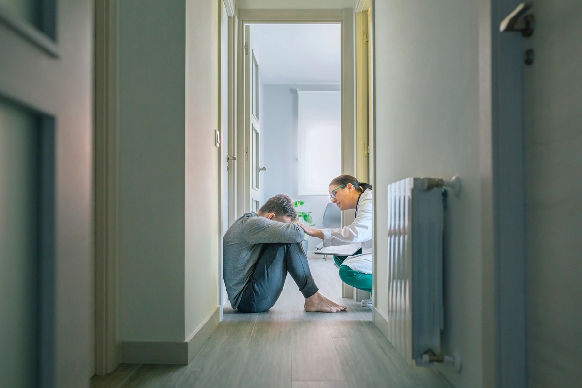 A man in inpatient care with his knees to his chest on a floor bing comforted by a nurse.