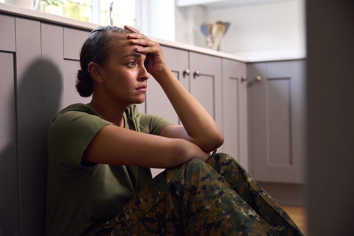 A woman in fatigues sits in front of her kitchen pantry, her hand on her forehead.