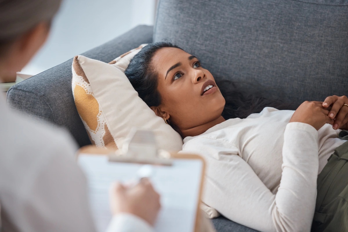 A woman in outpatient rehab lies on a couch, as her therapist takes notes on a clipboard in the foreground.