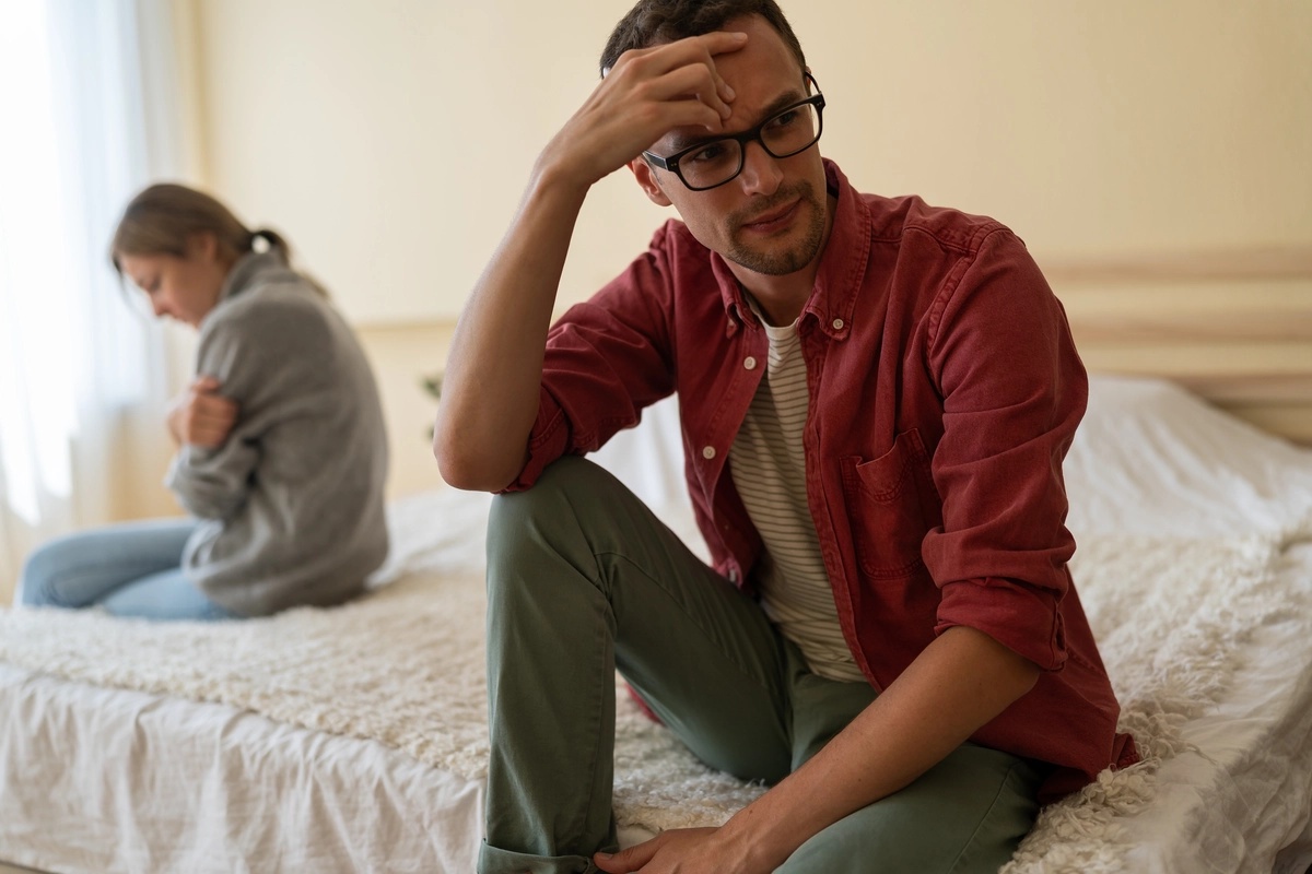 A man sits on a bed with his partner as he looks to the side. She looks depressed and is hugging herself.