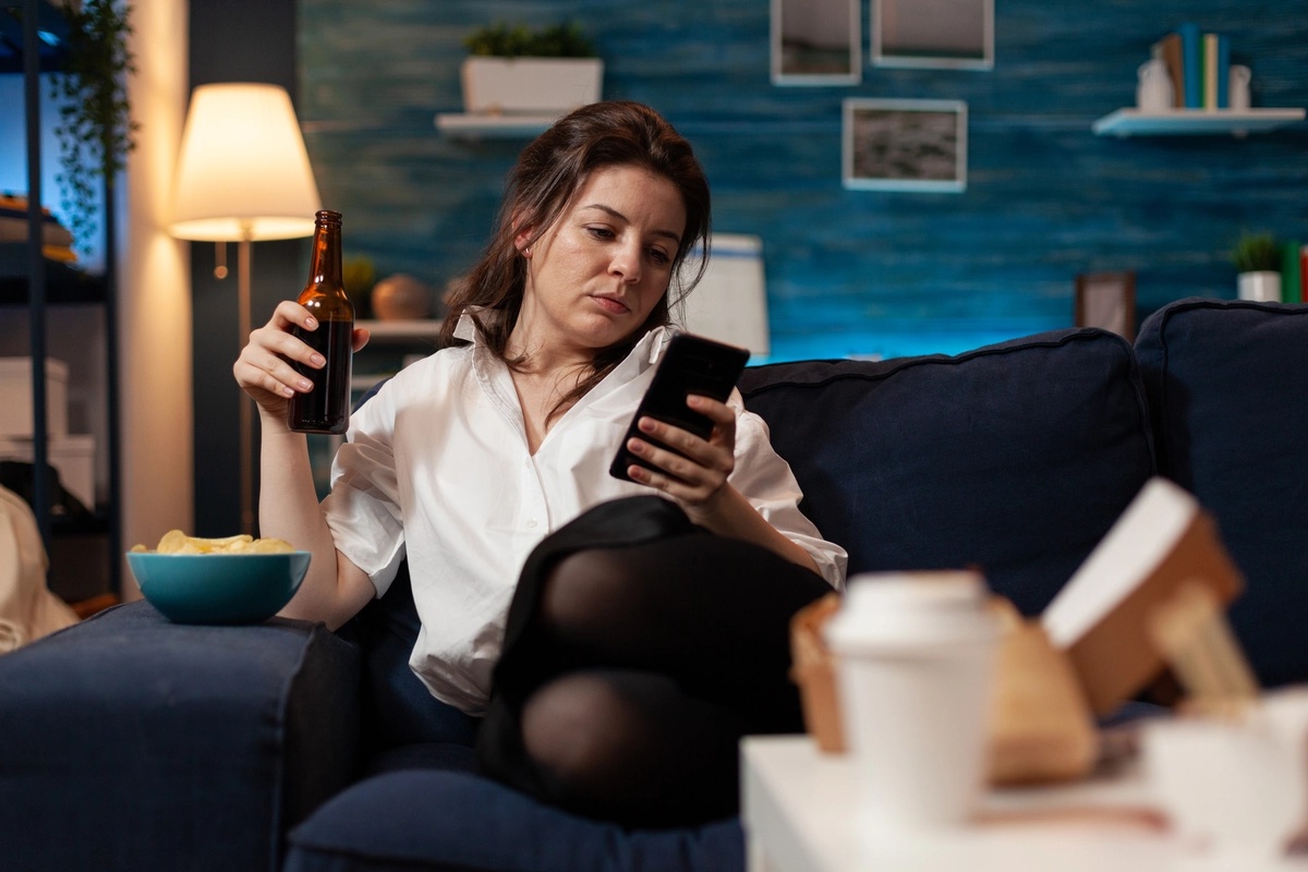 A woman with a phone addiction sitting on a couch and scrolling on her smartphone while sipping a beverage.