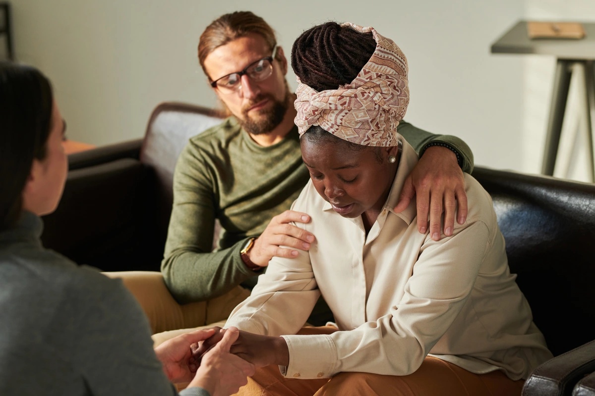 During a therapy session at an alcohol rehab, a man comforts a woman on a therapist's couch while the therapist looks on.