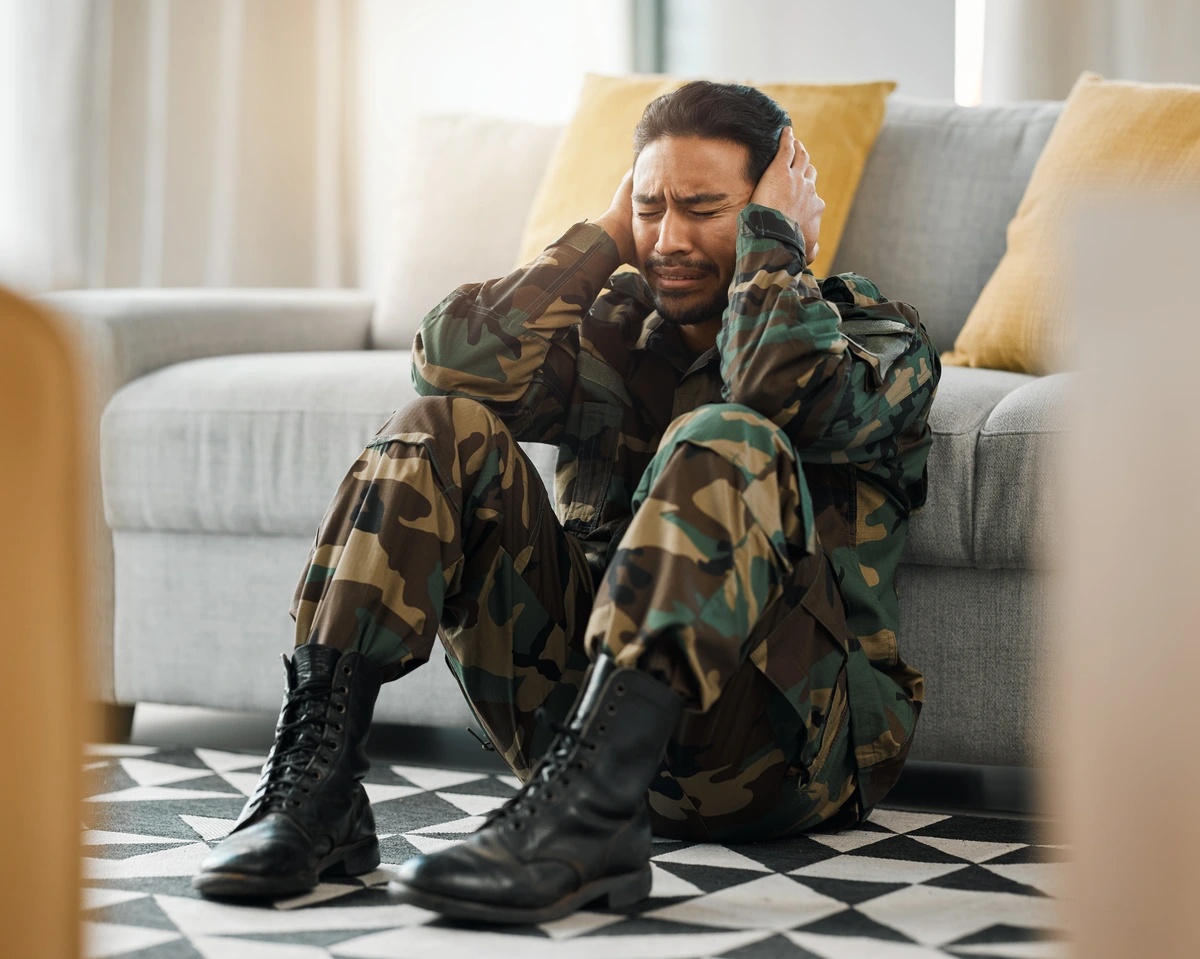 A man in camouflage sits on the floor in front of a sofa, suffering.