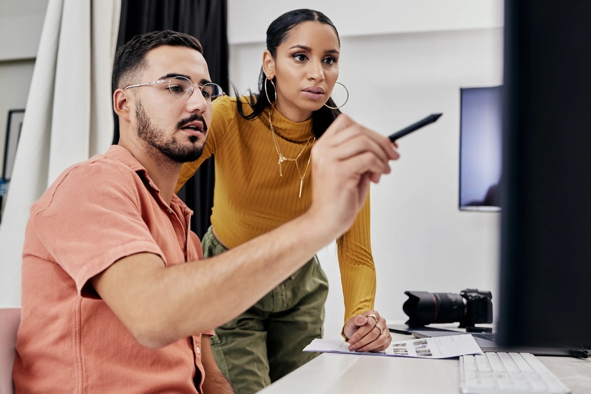 A man gestures at a monitor while a coworker watches.