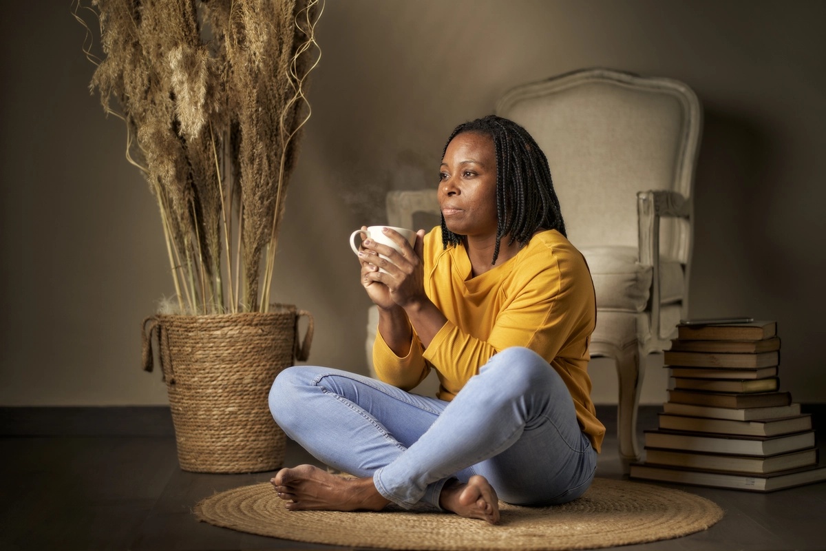 A woman sits crosslegged on a rug, with a cup of tea or coffee in her hands.