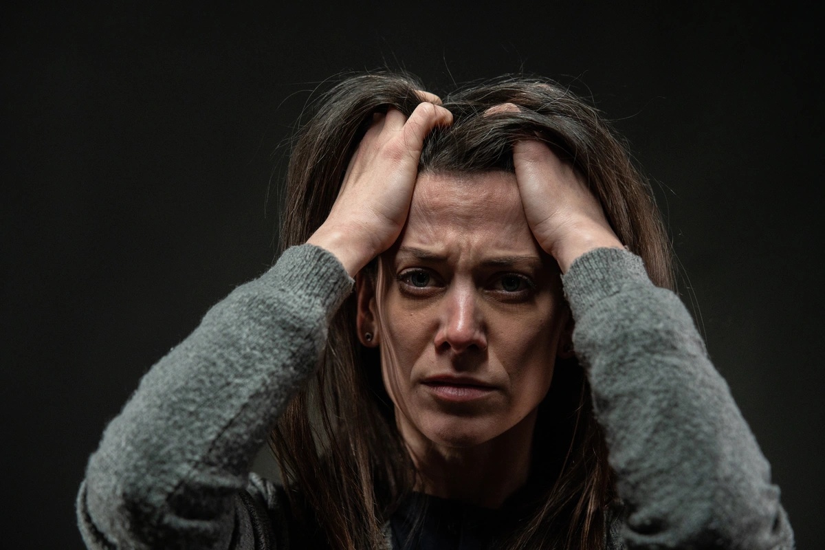 An anguished woman looks at the camera with her hands in her hair.