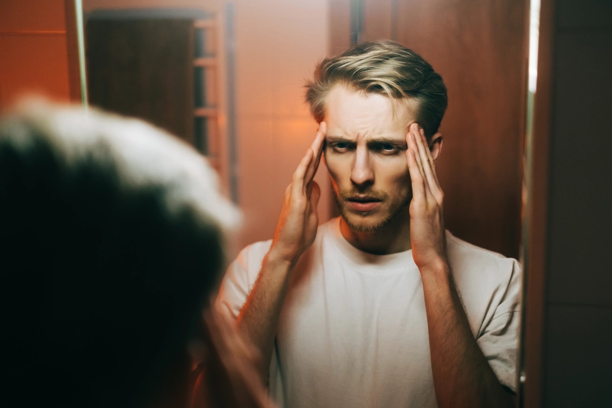 A man looking at himself in a mirror, holding his hands to his temples.