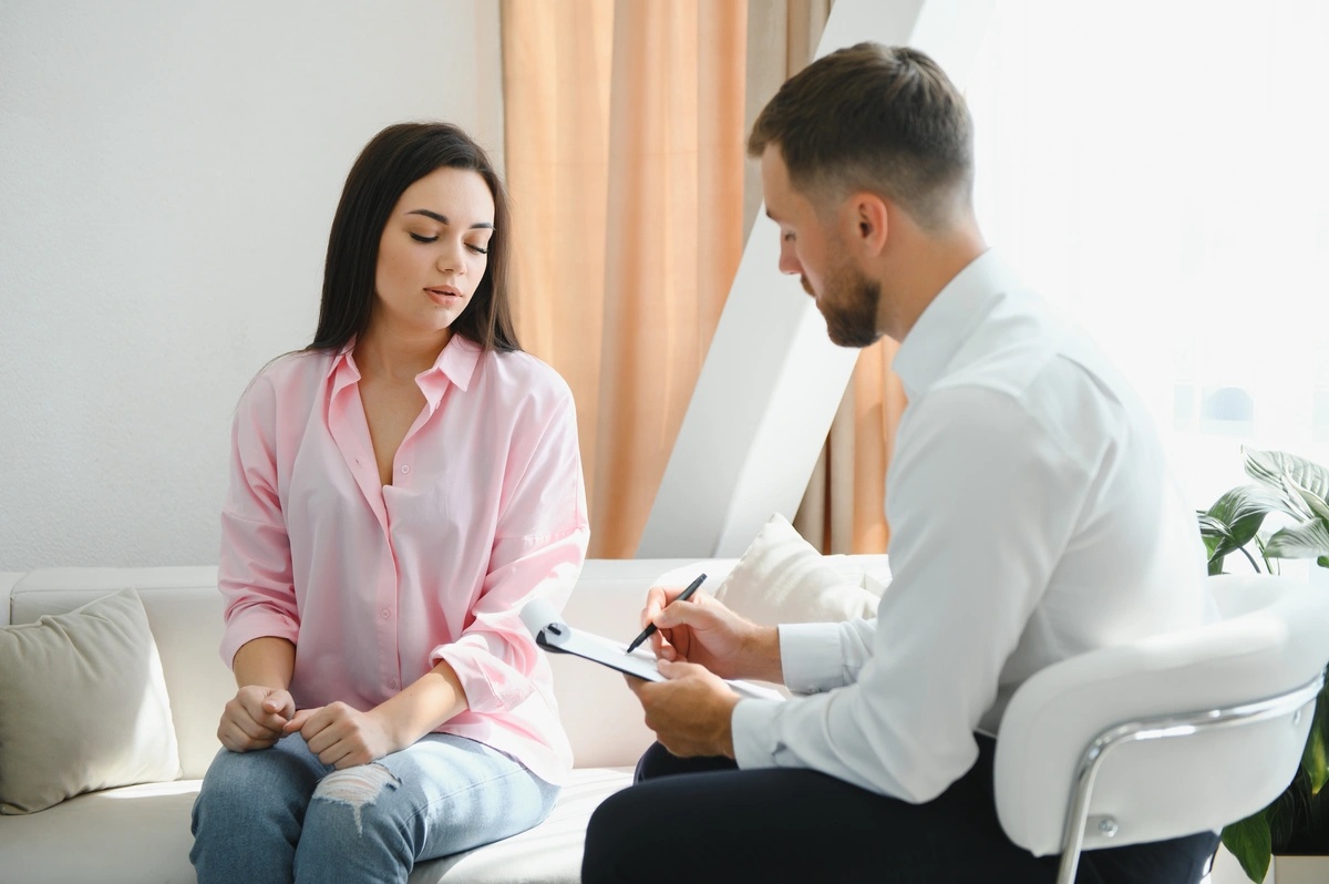 A woman sitting on a couch talks to a therapist, who is writing on a clipboard.