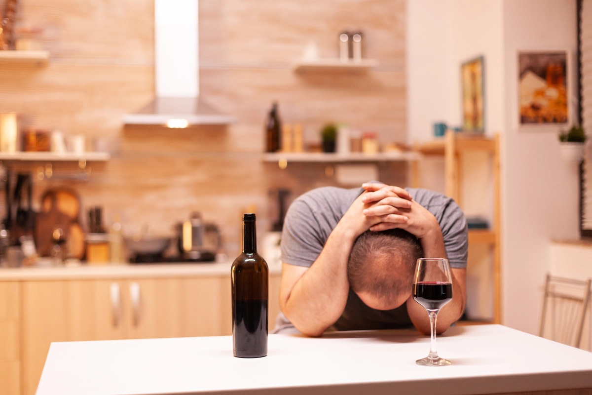 A man struggling with alcohol addiction sits at a table with a glass of wine and a bottle with his arms wrapped around his head.