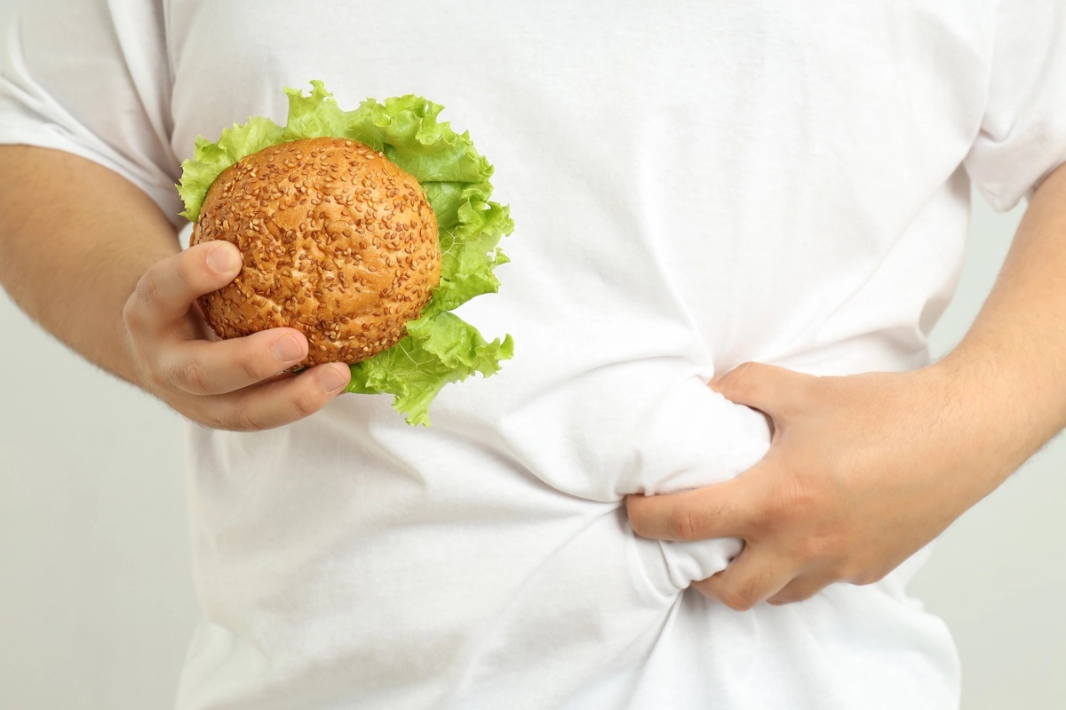 A man with a food addiction holding a burger in one hand and grabbing his stomach with the other hand.