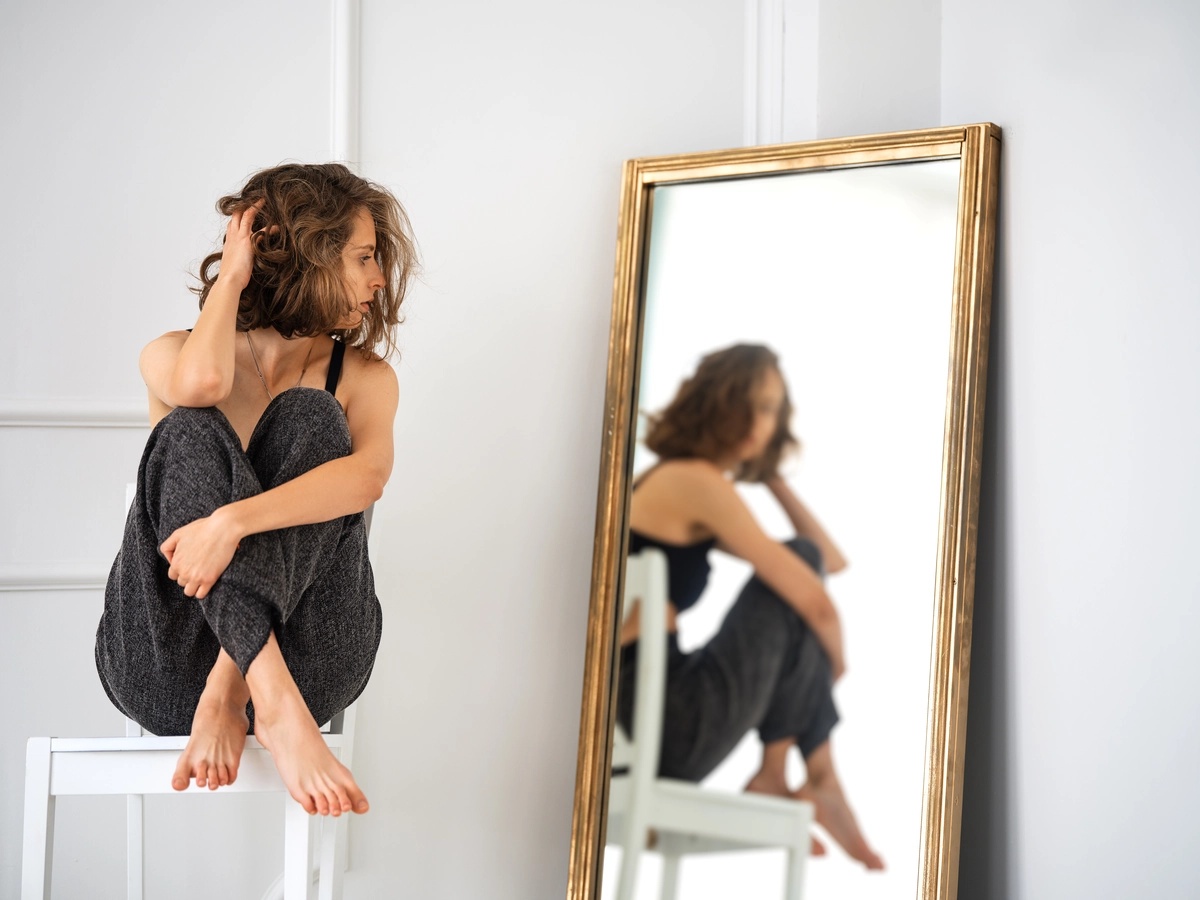 A woman sitting on a table stares at herself in a mirror.