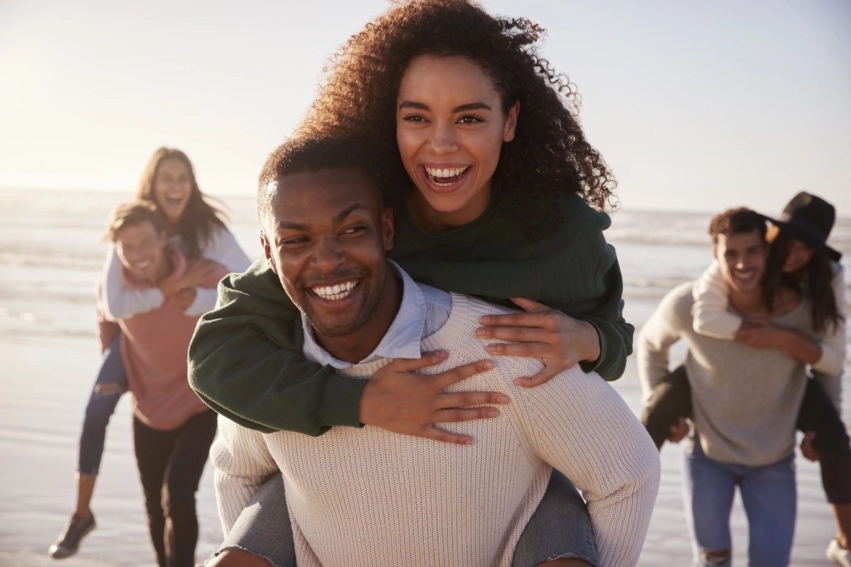 Drug rehab graduates celebrate on a beach, with a man carrying a woman on his back.