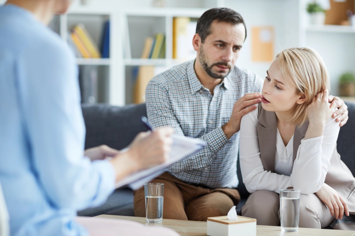 A man comforts a woman on a couch as a therapist takes notes.