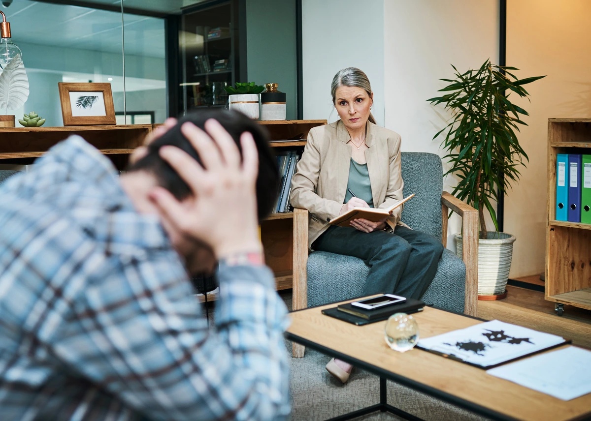 A therapist talks to her patient, who is holding his hands against his head.