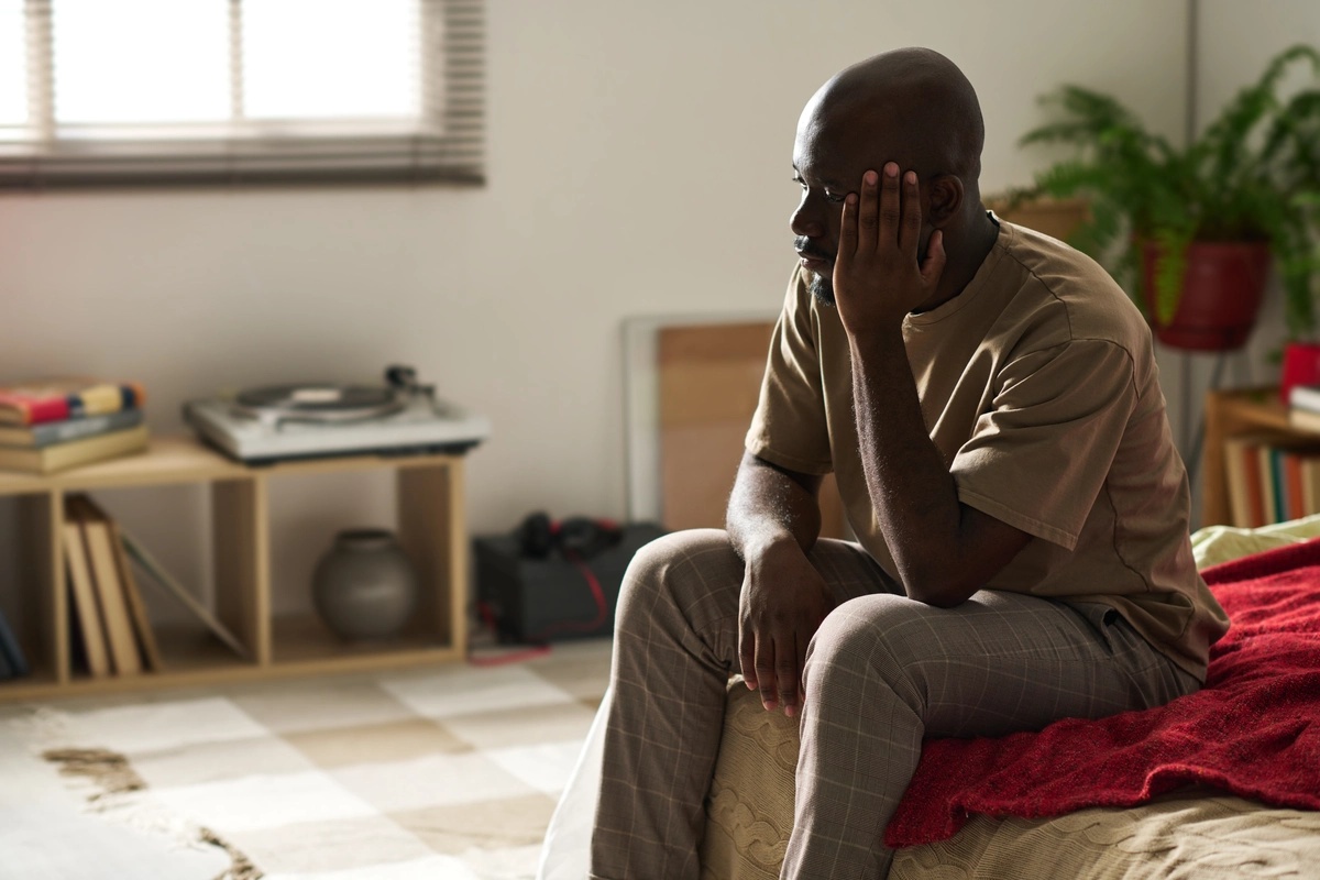 A man sitting on the edge of his bed with his hand up to his face in a sad manner, possibly struggling with symptoms of persistent depressive disorder.