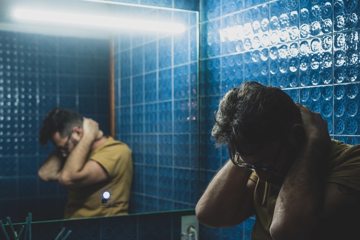 A man leans against a bathroom wall with his hands on his neck.