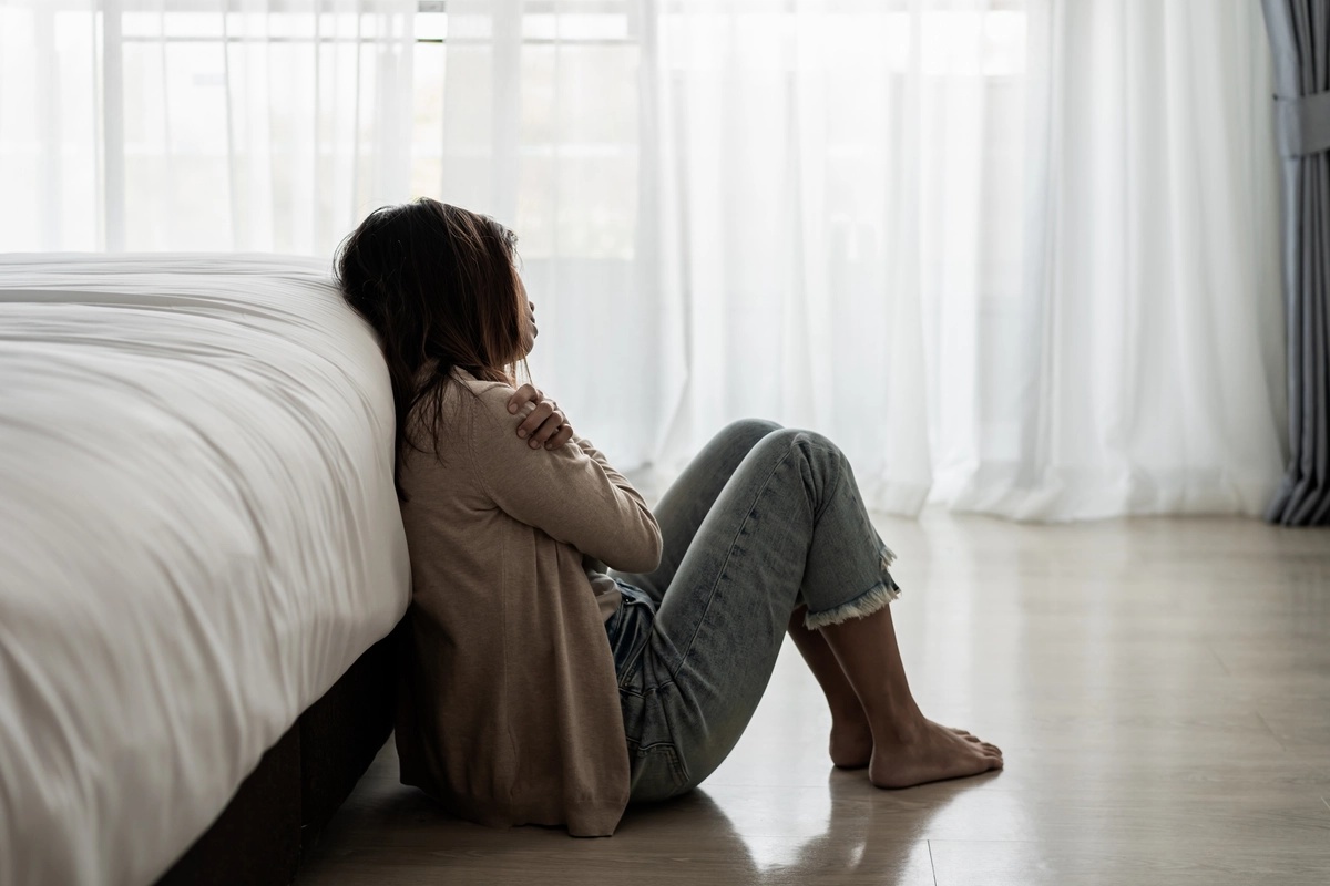 Woman sitting on the floor next to her bed.