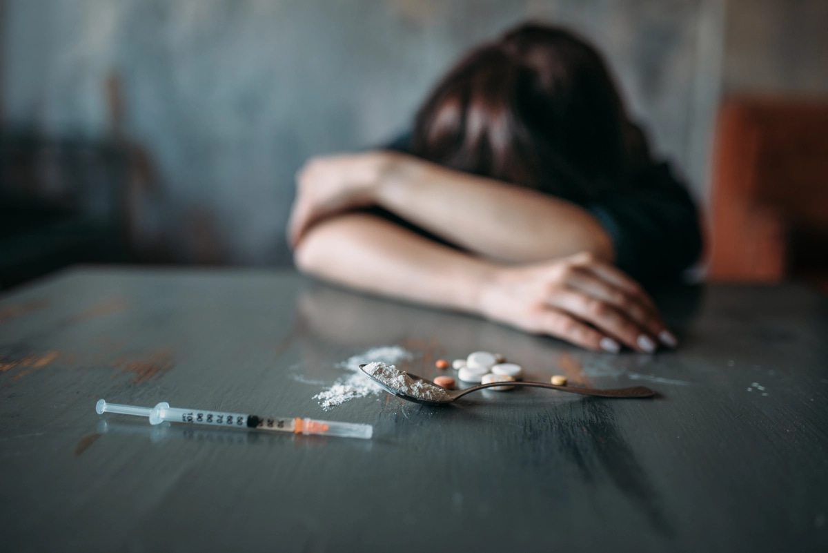 A person struggling with meth addiction lays her head on her arms at a table, with a needle and meth also on the table.